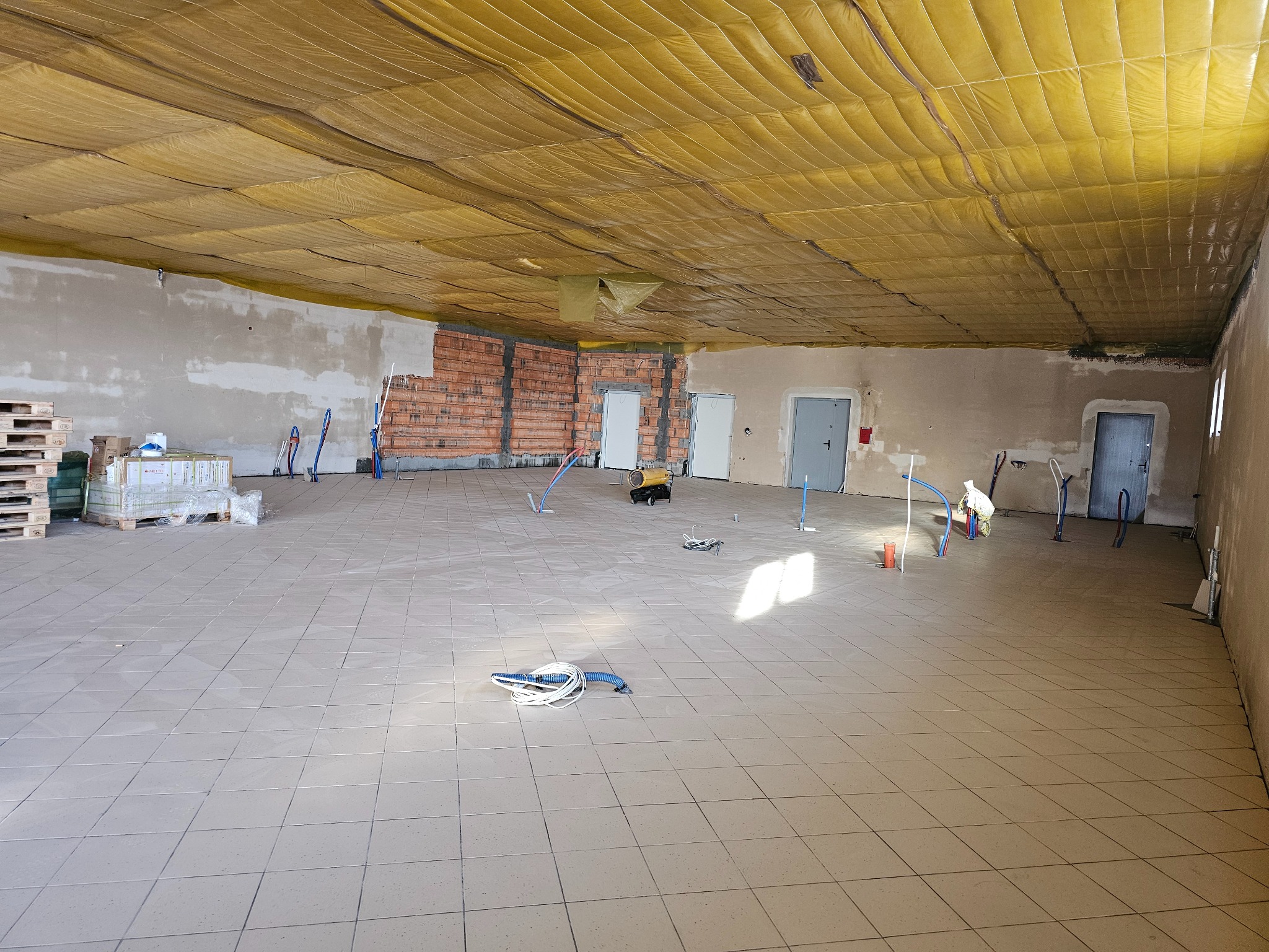 Duże pomieszczenie w trakcie prac wykończeniowych: widoczne ocieplenie sufitu folią, surowa ściana z cegły, częściowo położone płytki na podłodze oraz wystające rury instalacji.