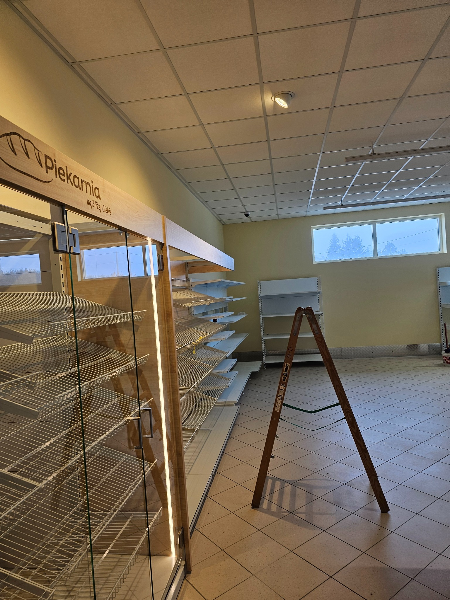 Wnętrze piekarni w trakcie prac wykończeniowych: drewniany regał z metalowymi półkami, drabina oparta o ścianę, puste białe regały w tle, widoczne okno z zewnątrz.