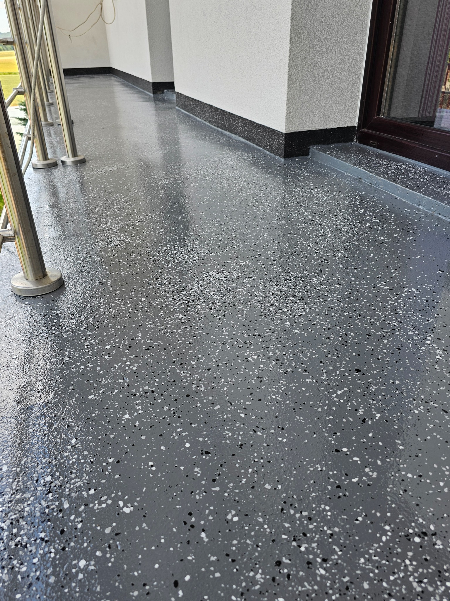 Błyszcząca, szara posadzka z żywicy epoksydowej na balkonie, wykończona czarnym cokolikiem przy ścianie i metalową balustradą.
