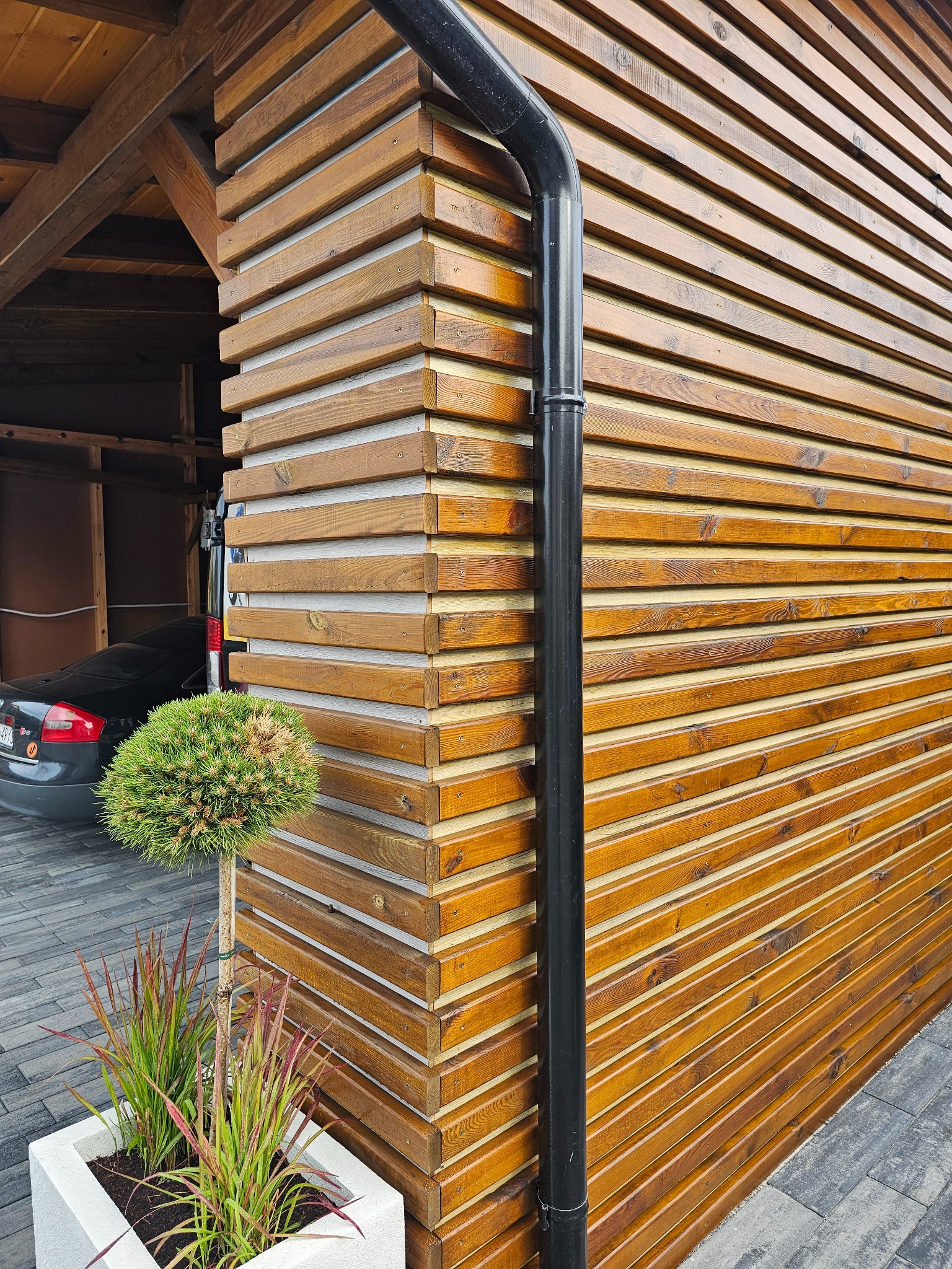 Drewniana elewacja budynku z poziomych desek, czarna rynna, kostka brukowa i dekoracyjne rośliny w donicy. W tle widoczny samochód.