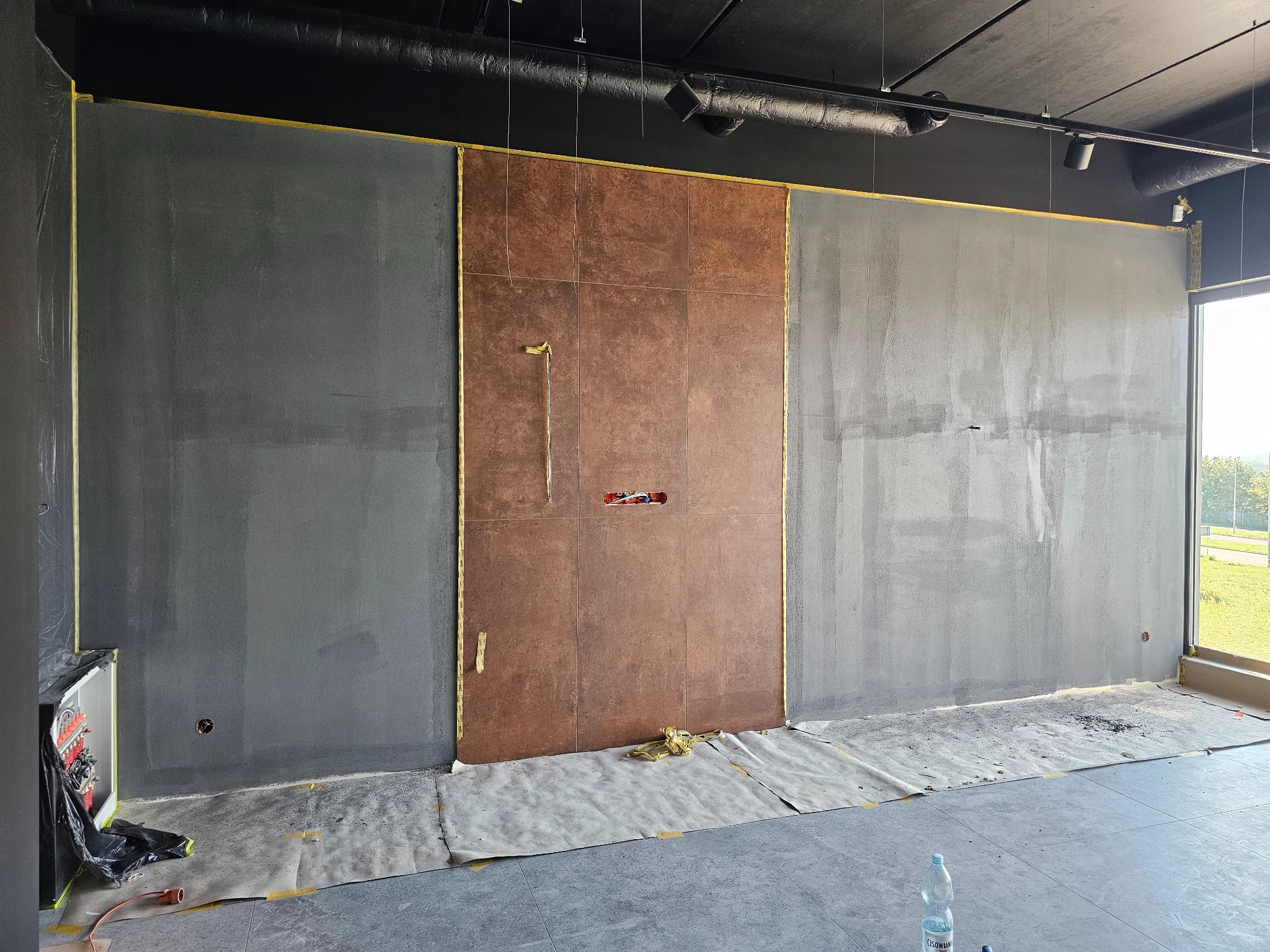Ściana w trakcie prac wykończeniowych: szare ściany po bokach, centralnie brązowe płytki, zabezpieczona podłoga, widoczne instalacje elektryczne.