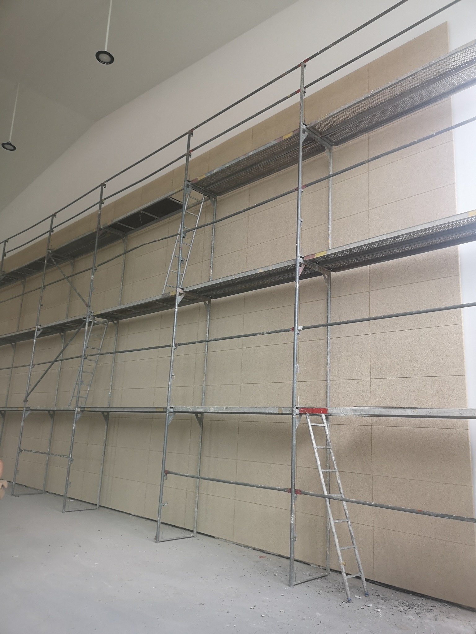 Montaż płyt na ścianie z użyciem rusztowania. Widoczne płyty w odcieniach beżu, metalowe rusztowanie i drabina. Oświetlenie sufitowe.