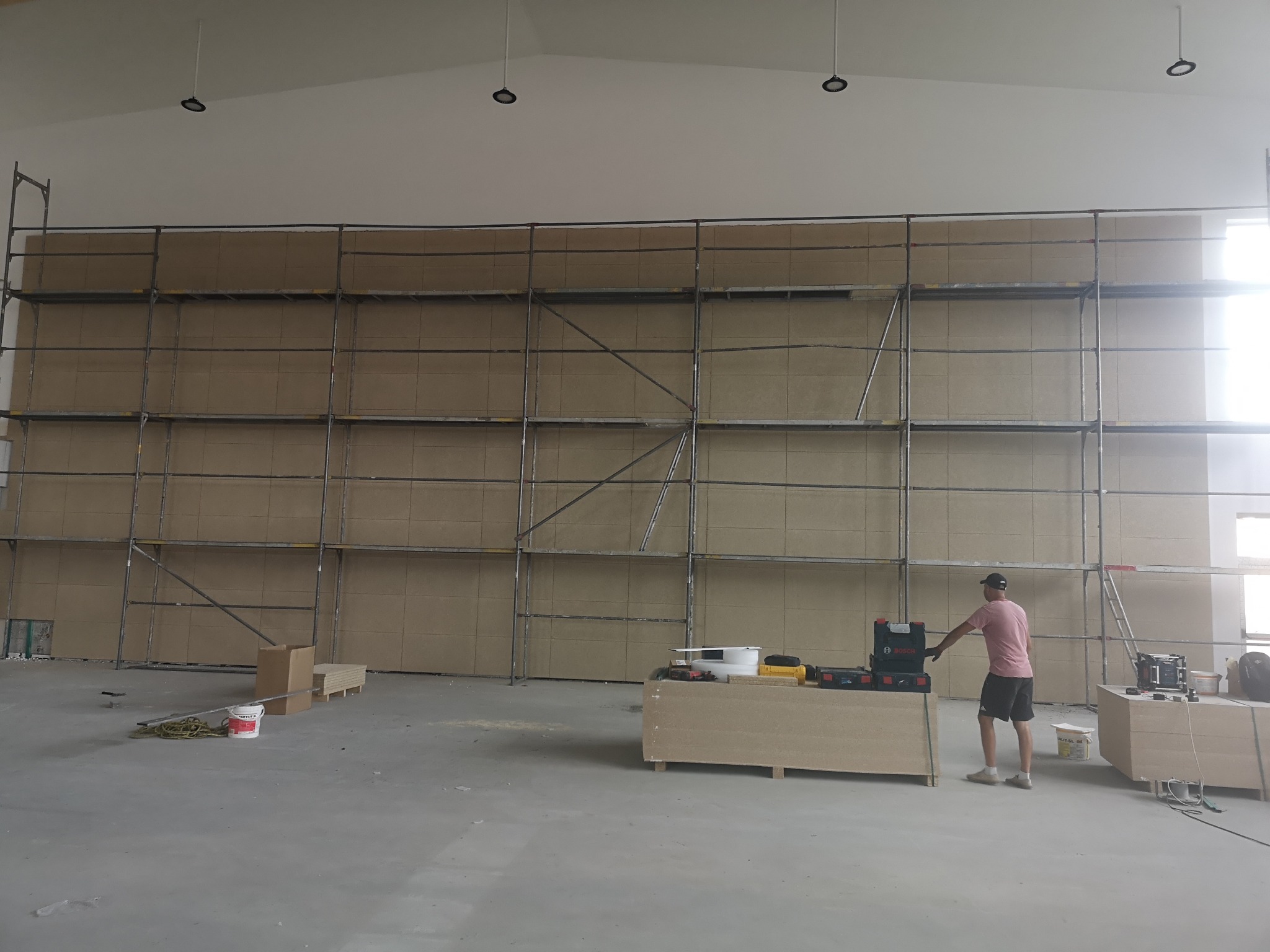 Montaż płyt na ścianie w dużym pomieszczeniu. Widoczne rusztowania, narzędzia i pracownik w trakcie pracy. Oświetlenie sufitowe.