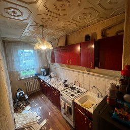 Andrzej Dziuk - Kuchenne wnętrze w trakcie remontu: zerwane płyty sufitowe leżą na podłodze, widoczna drabina, ściany przygotowane do odświeżenia, meble kuchenne w kolorze czerwonym, stare okno z firanką.