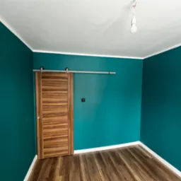 Pomalowane na turkusowo wnętrze pokoju z drewnianą przesuwną szafą, widoczne panele podłogowe i białe listwy przypodłogowe.