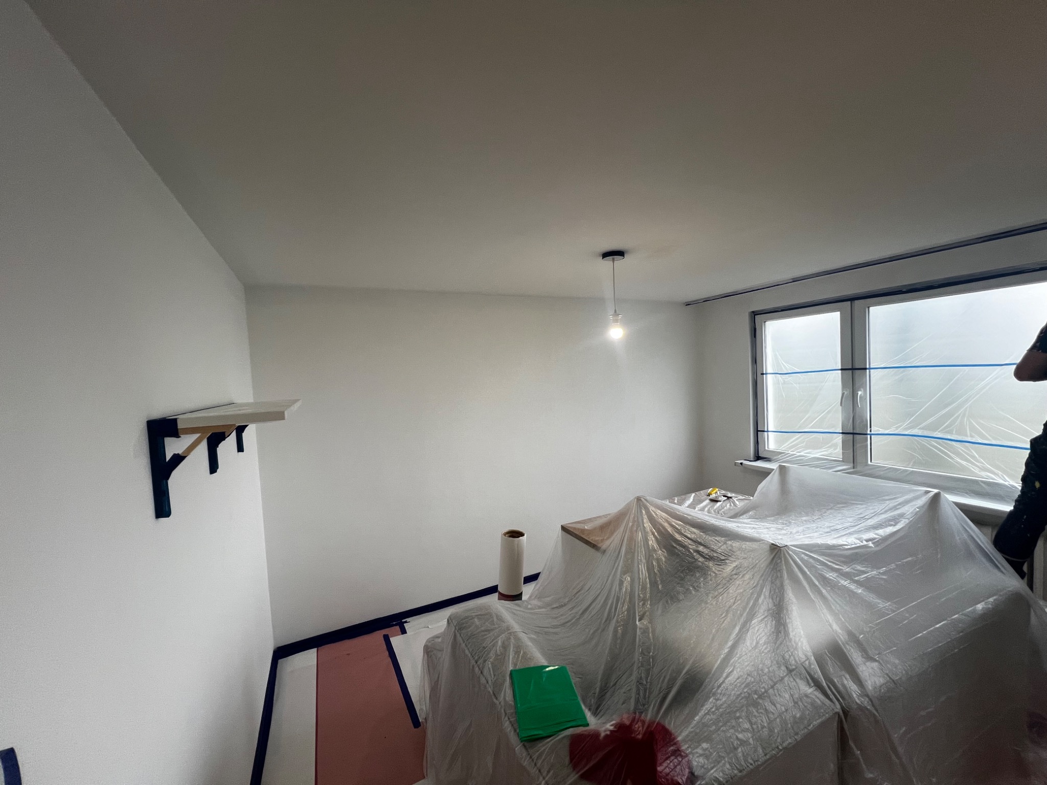 Białe ściany w pokoju zabezpieczonym folią malarską i taśmą, przygotowanym do malowania; widoczna półka na ścianie, rolka folii i lampa sufitowa.