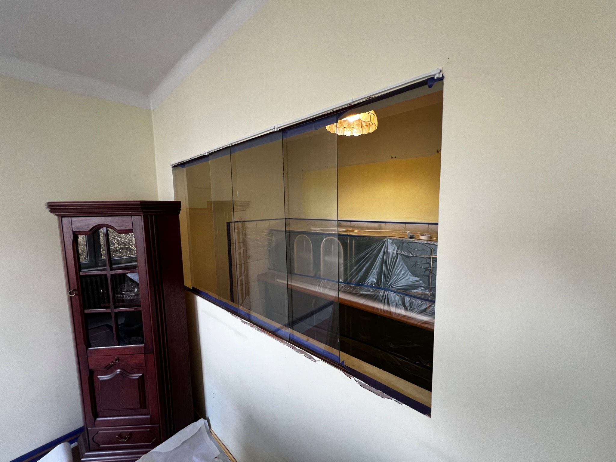 Przygotowania do malowania: ściana zabezpieczona folią i taśmą malarską, widoczna część mebli przykrytych folią ochronną, brązowa witryna w rogu pokoju.
