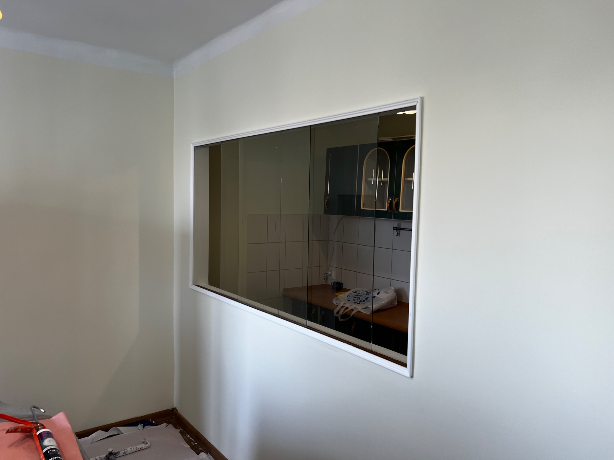 Świeżo pomalowana ściana z białym obramowaniem otworu, widoczne narzędzia malarskie przy podłodze, wnętrze kuchni widoczne przez otwór.