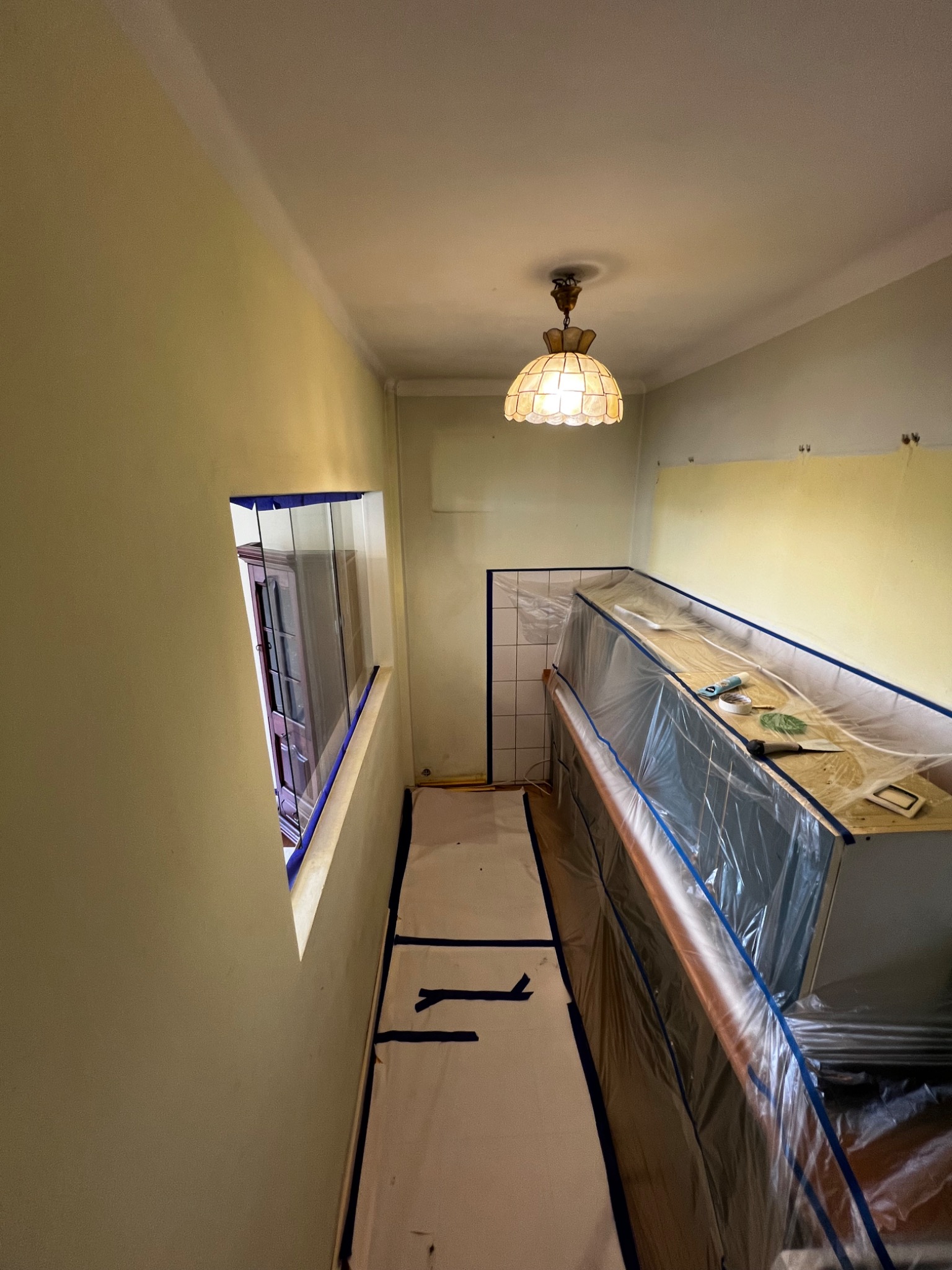 Przygotowanie do malowania korytarza w Sosnowcu: zabezpieczone folią meble i podłoga, oklejone okno, widoczna lampa sufitowa i fragment pomalowanej ściany.