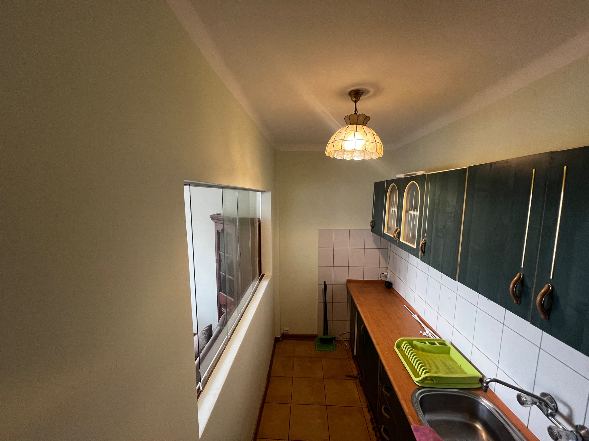 Kuchnia w mieszkaniu z zielonymi szafkami, brązowym blatem i beżowymi ścianami, widok na salon przez okno, oświetlona lampą z kloszem w stylu retro.