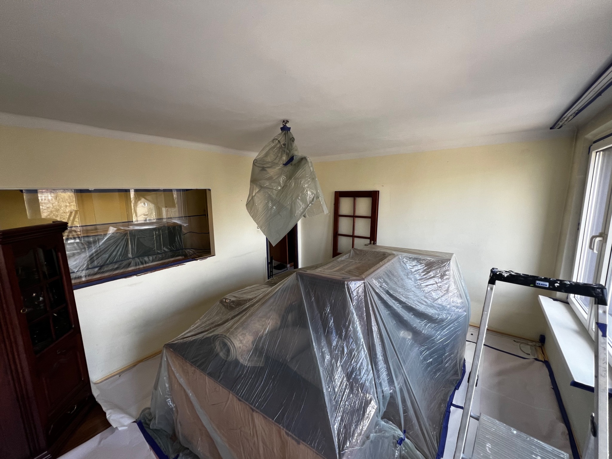 Pokój przygotowany do prac malarskich: meble i żyrandol zabezpieczone folią, ściany z taśmą malarską, drabina oparta o parapet, widoczna szafka z ciemnego drewna.