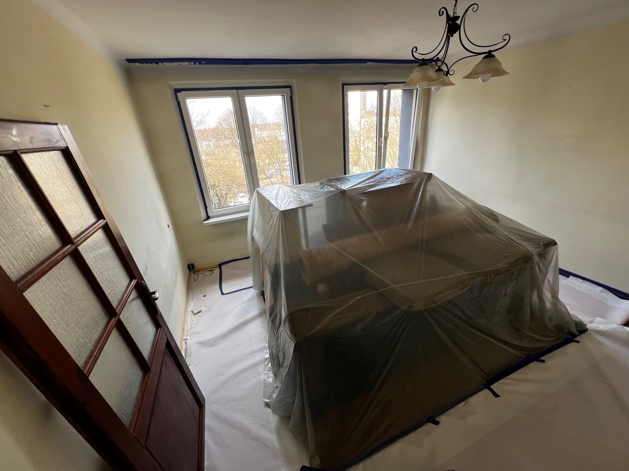 Pokój przygotowany do prac malarskich: zabezpieczone okna, listwy i meble folią ochronną, ściany w odcieniach beżu, widoczny żyrandol.