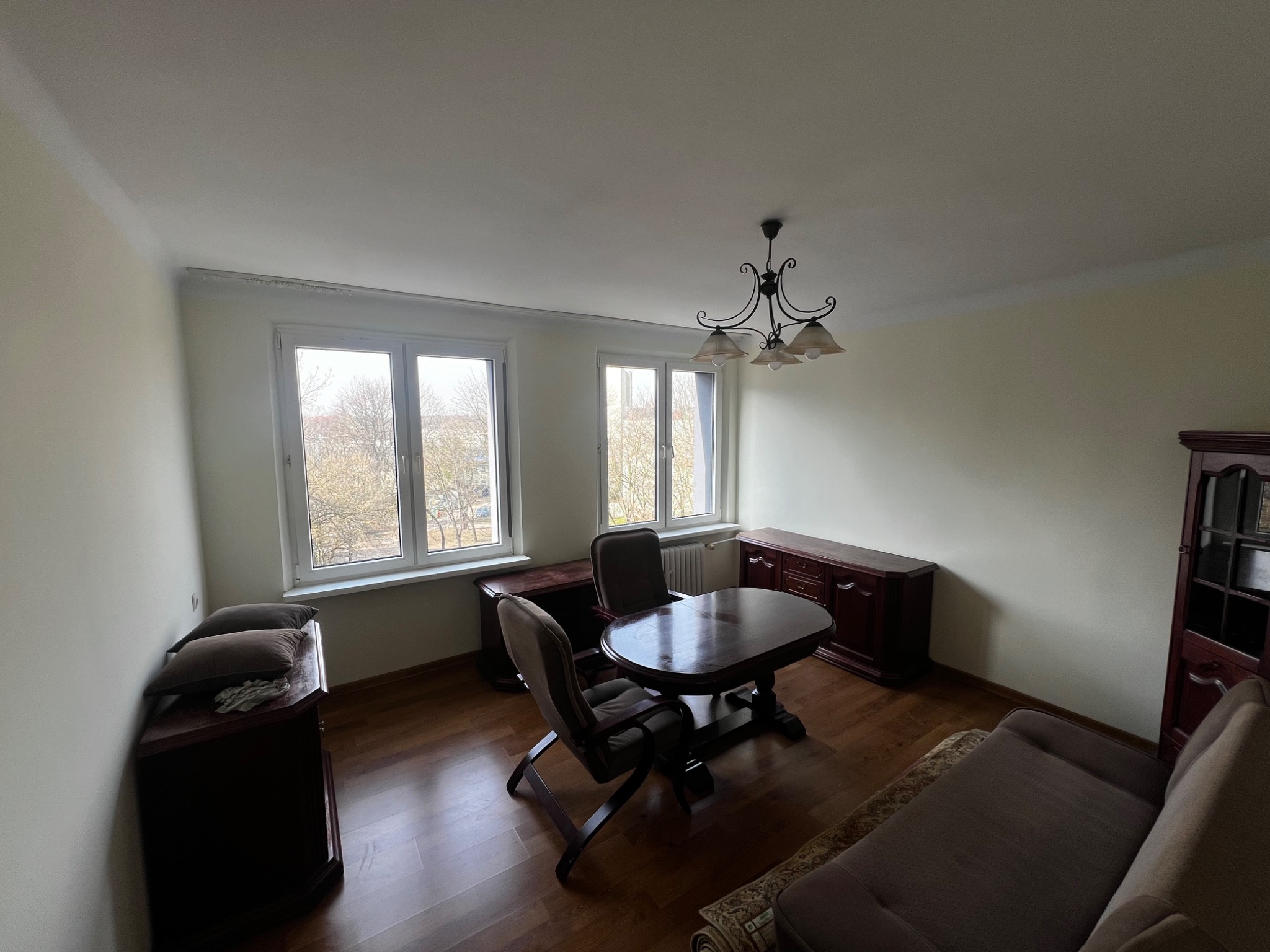 Jasne wnętrze pokoju z parkietem, brązowymi meblami i widokiem z okna na drzewa bez liści. Ściany w odcieniach kremu, oświetlenie centralne z żyrandolem.