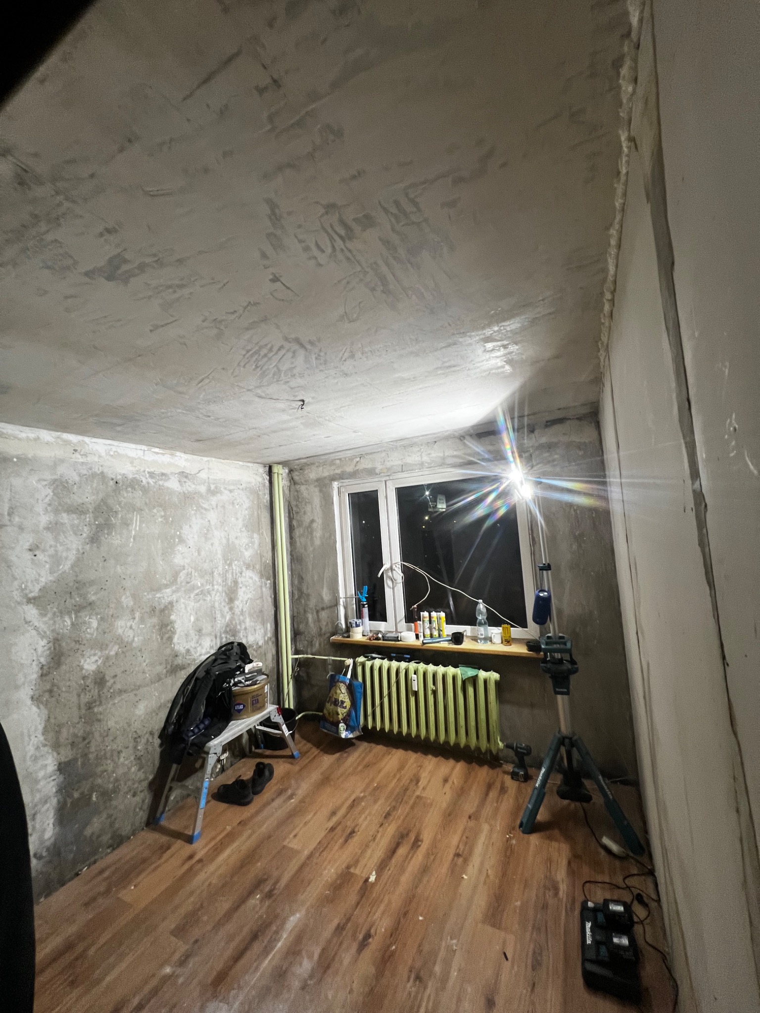 Pokój w trakcie remontu: surowa ściana z widoczną strukturą, częściowo wygładzony sufit, ułożone panele podłogowe, grzejnik pod oknem i lampa budowlana na statywie.