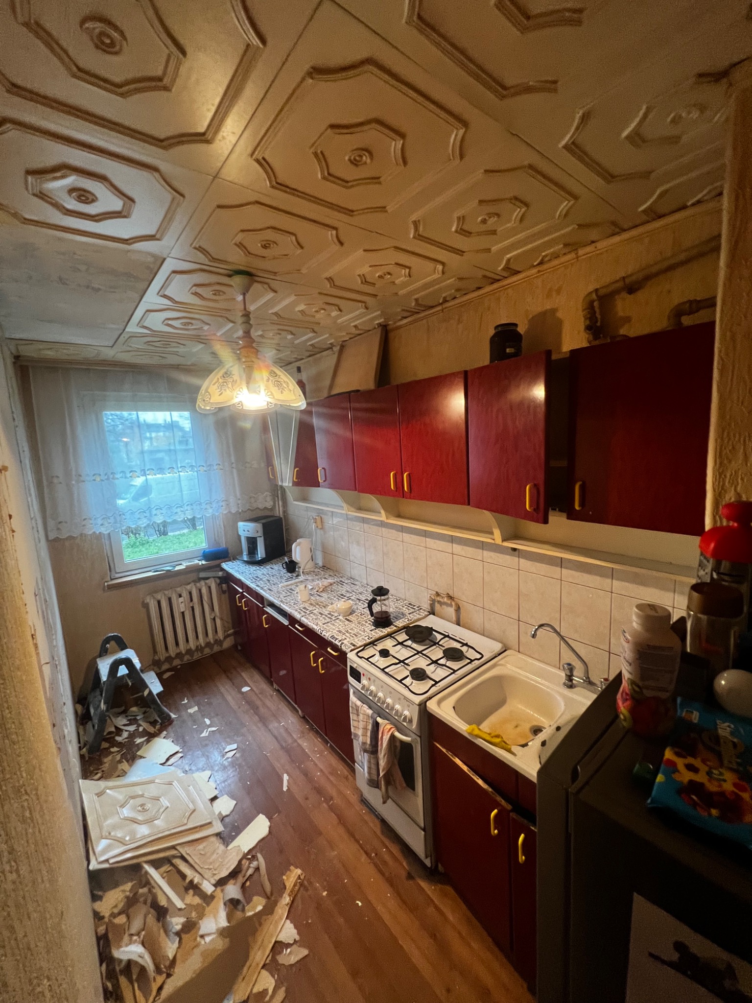 Kuchenne wnętrze w trakcie remontu: zerwane płyty sufitowe leżą na podłodze, widoczna drabina, ściany przygotowane do odświeżenia, meble kuchenne w kolorze czerwonym, stare okno z firanką.