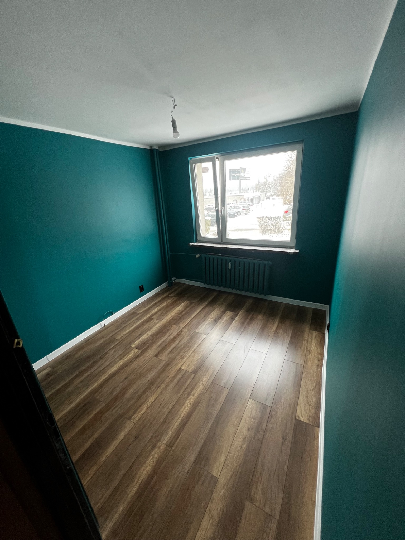 Pomalowane na butelkową zieleń ściany i ułożone panele podłogowe w pokoju w Sosnowcu, widok na okno z grzejnikiem i zimowy krajobraz za szybą.