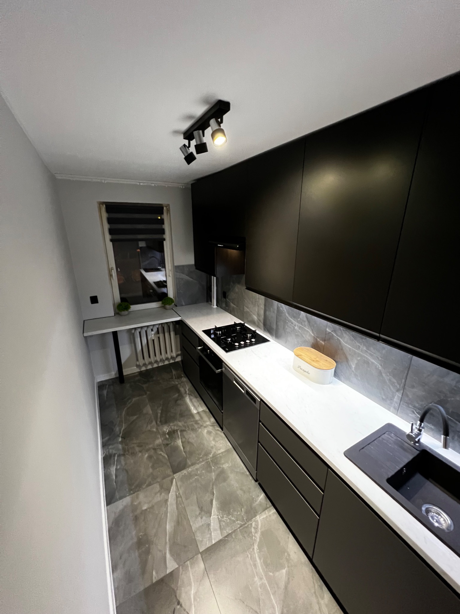 Nowoczesna, wąska kuchnia w szaro-czarnej tonacji z marmurową podłogą i białym blatem, oświetlona reflektorami sufitowymi.