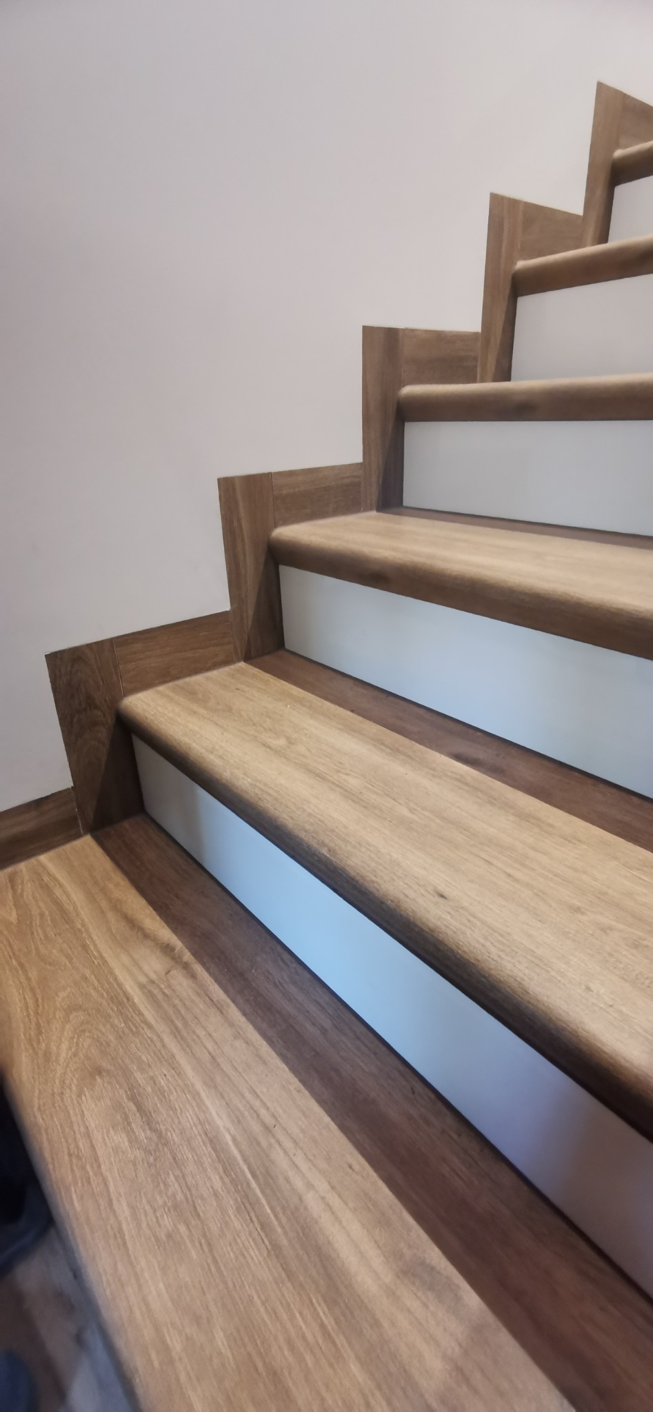 Schody z drewnianymi stopniami i białymi podstopnicami, wykończone listwami przypodłogowymi w kolorze drewna, widok z dołu.