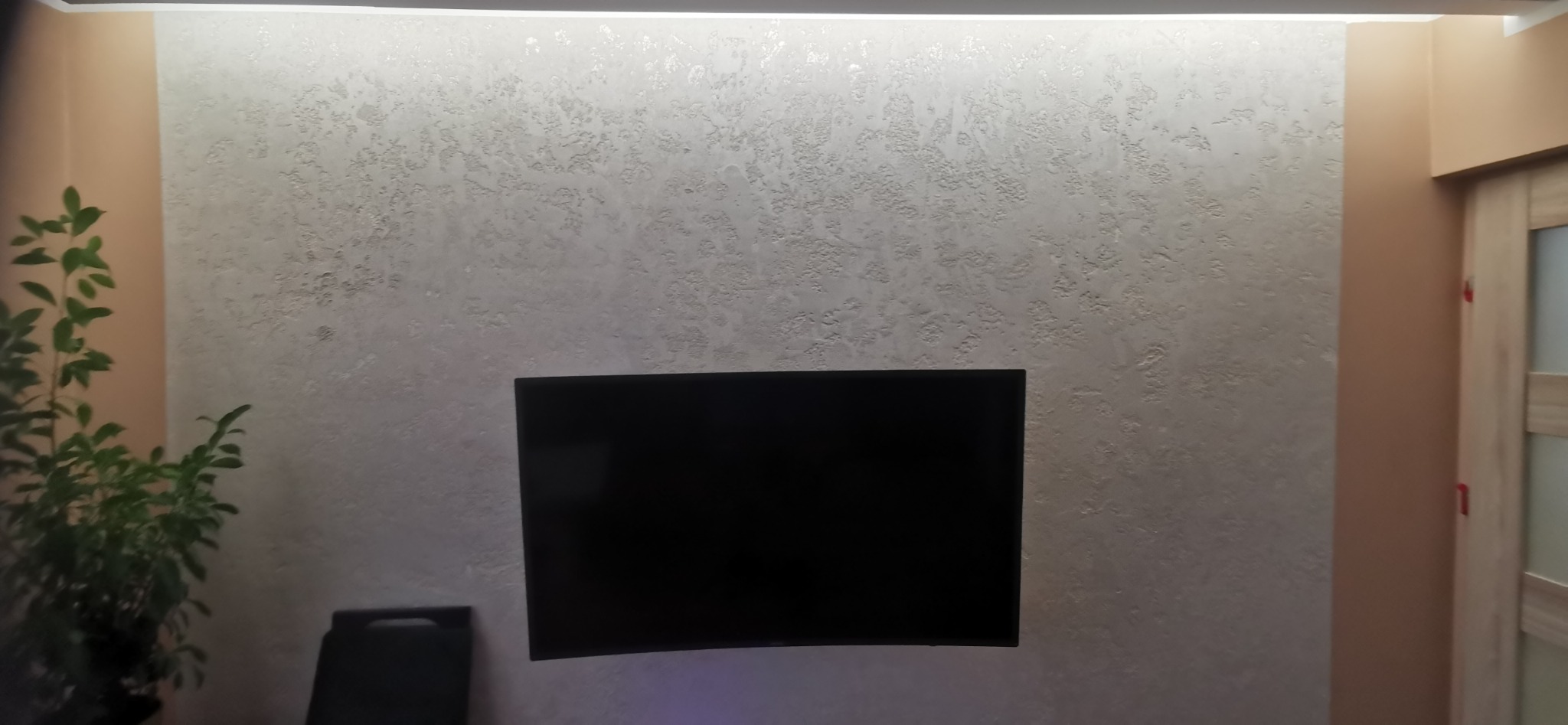 Ściana z dekoracyjną, jasną gładzią strukturalną podświetloną od góry, z zawieszonym telewizorem i rośliną doniczkową po lewej stronie.