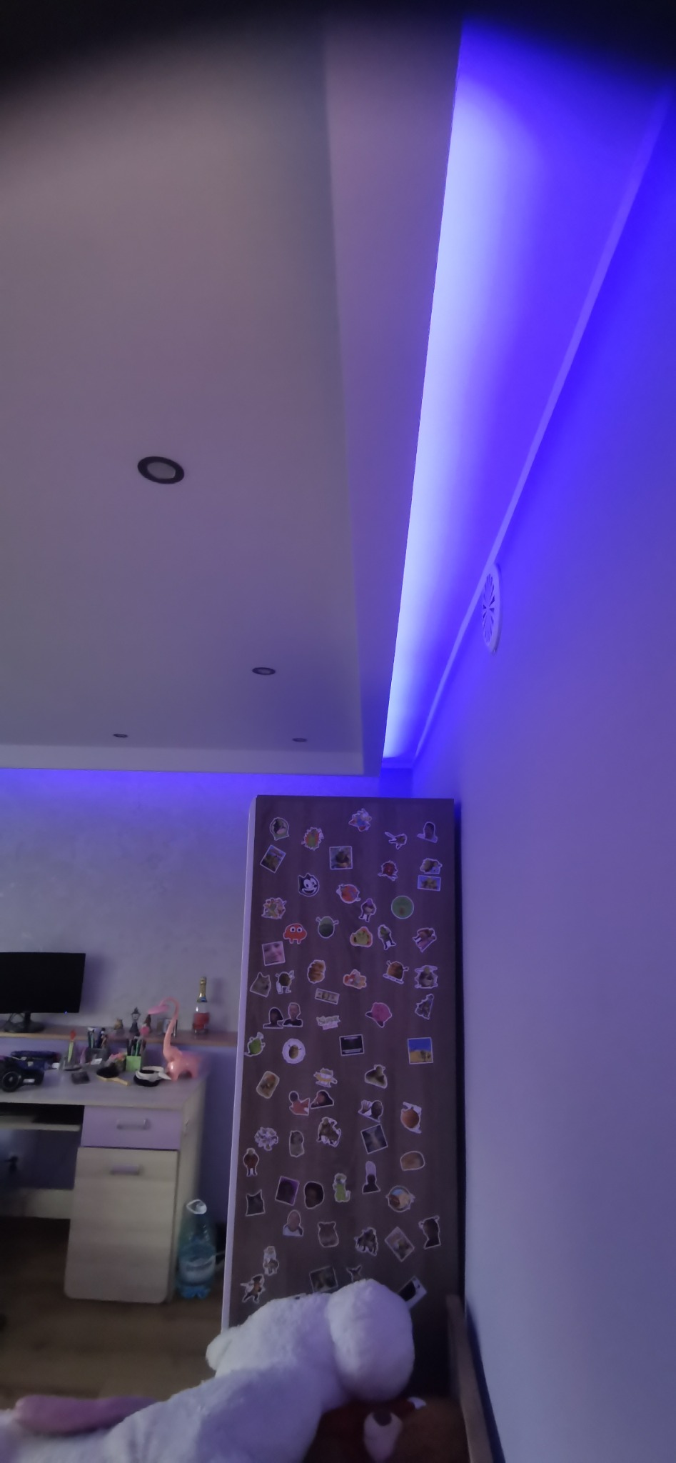 Pokój z podwieszanym sufitem i oświetleniem LED w kolorze niebieskim, widoczny fragment biurka z akcesoriami i lodówka oklejona naklejkami.