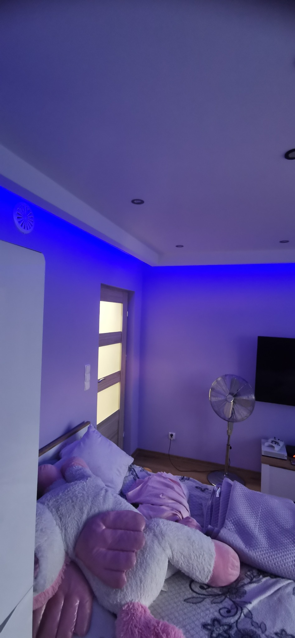 Pokój z niebieskim oświetleniem LED na suficie, widoczny wentylator stojący, łóżko z pluszowym jednorożcem i telewizor na ścianie.