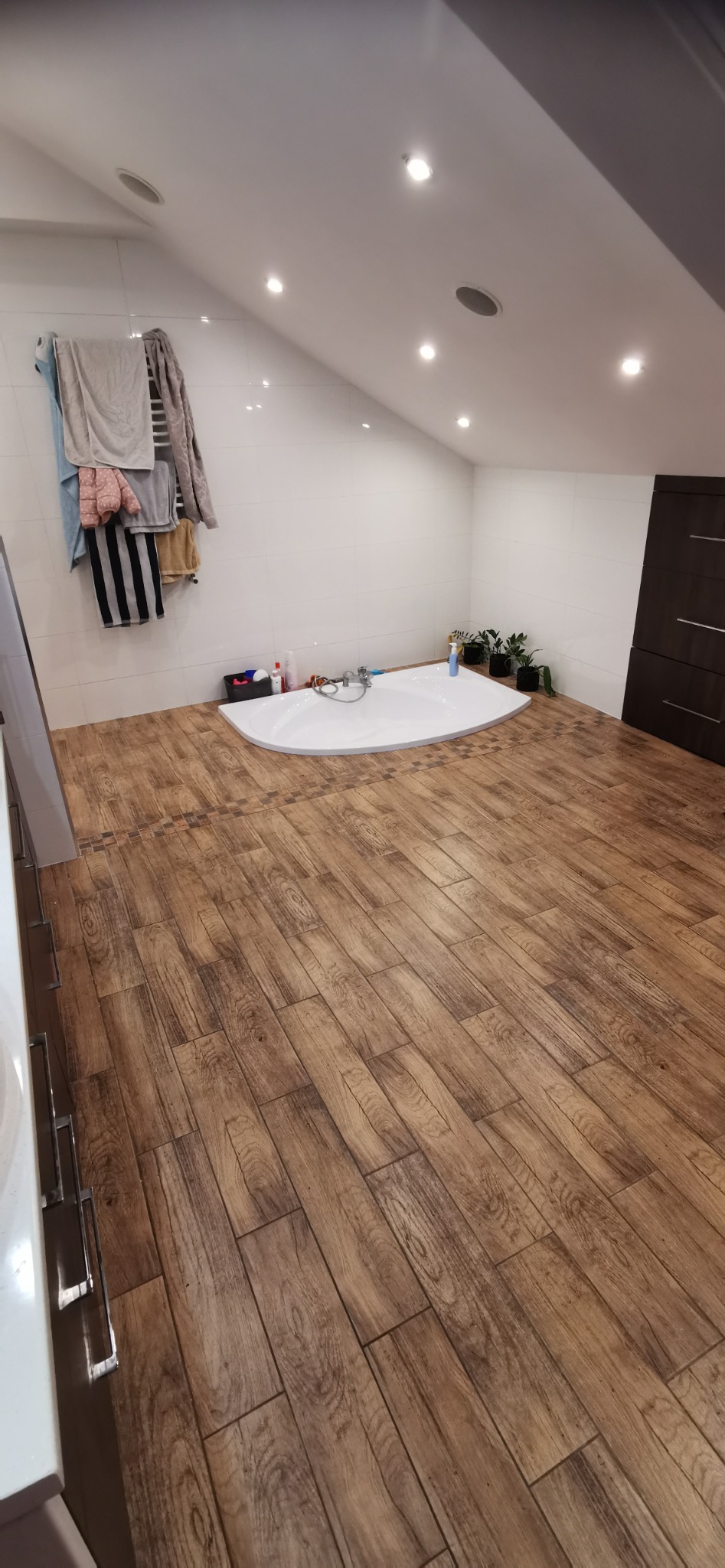 Ukończona łazienka na poddaszu z drewnopodobną podłogą z paneli, narożną białą wanną, białymi płytkami na ścianach i suszarką z ręcznikami.