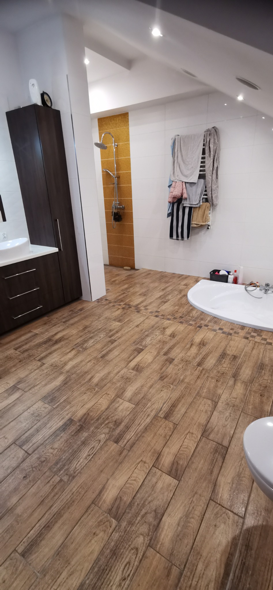 Wykończona łazienka na poddaszu z drewnopodobną podłogą ułożoną w cegiełkę, kabiną prysznicową z panelami w odcieniach brązu, białą wanną narożną i szafką z umywalką.