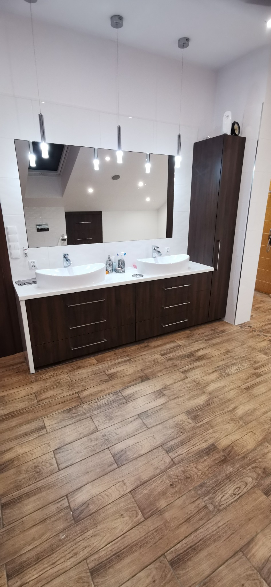 Nowoczesna łazienka z podwójną umywalką nablatową, szafką z ciemnego drewna i dużym lustrem z oświetleniem punktowym, uzupełniona podłogą z płytek imitujących drewno.