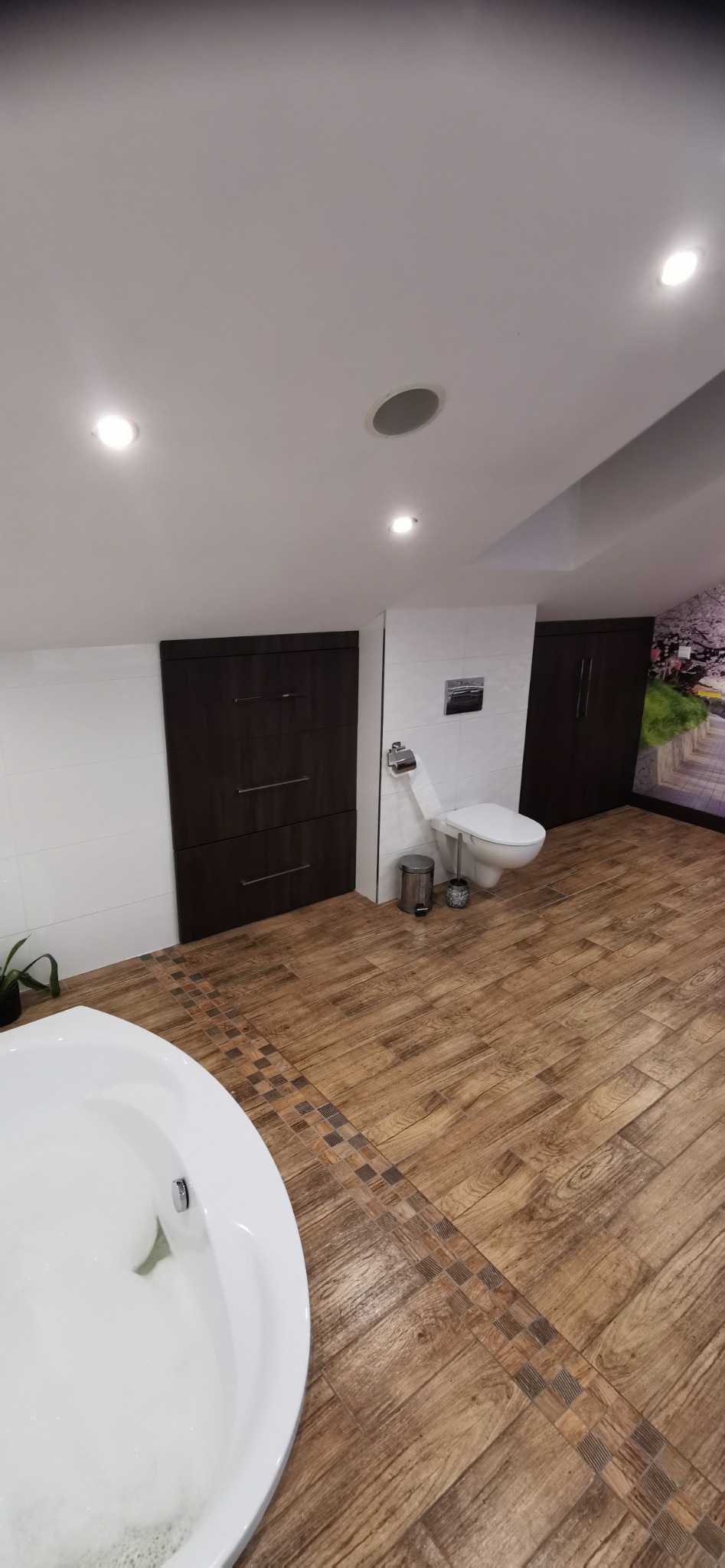 Nowoczesna łazienka na poddaszu z drewnopodobną podłogą, białą wanną wypełnioną pianą, toaletą, zabudową meblową w ciemnym brązie oraz dekoracyjną fototapetą z motywem krajobrazowym.