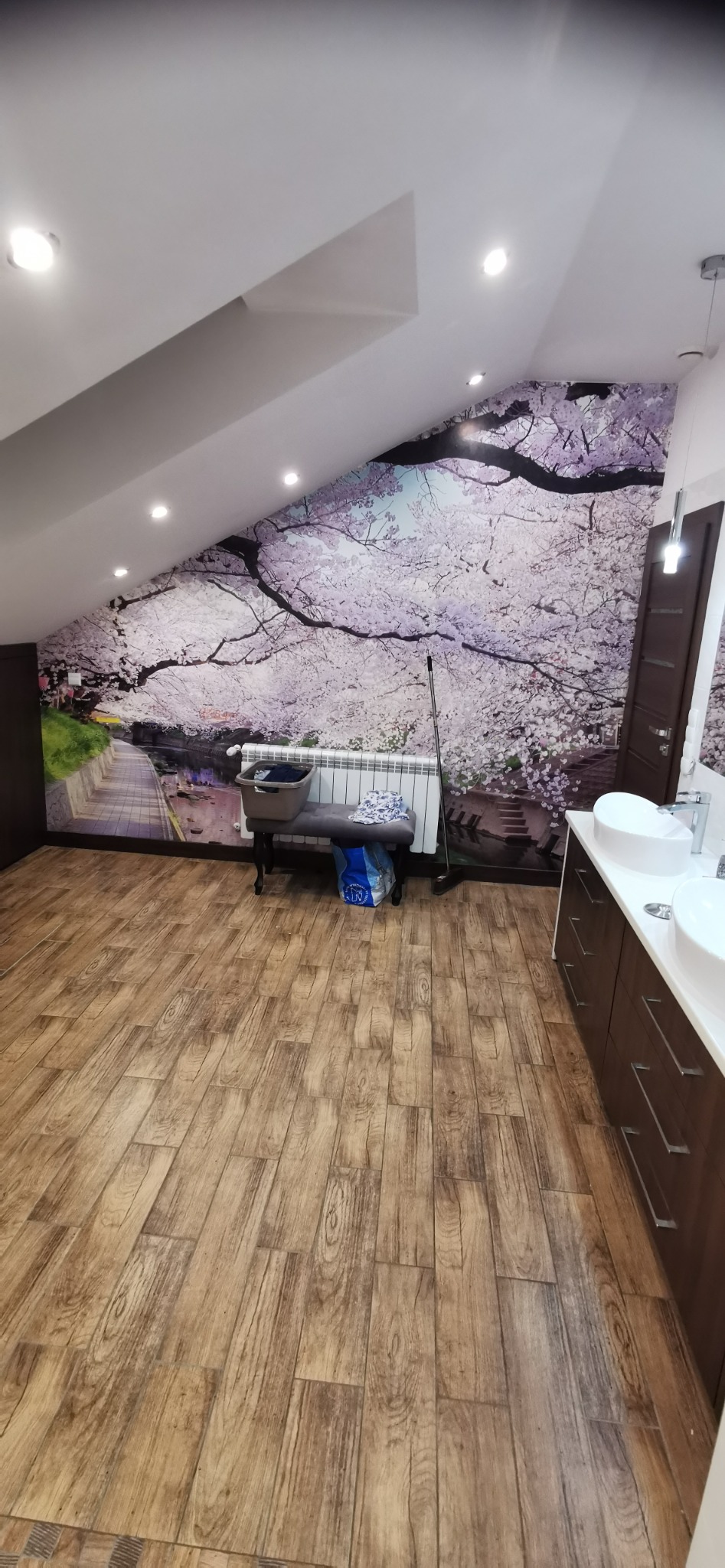 Łazienka na poddaszu z drewnopodobną podłogą, umywalkami nablatowymi na ciemnych szafkach i fototapetą z motywem kwitnącej wiśni na ścianie skośnej