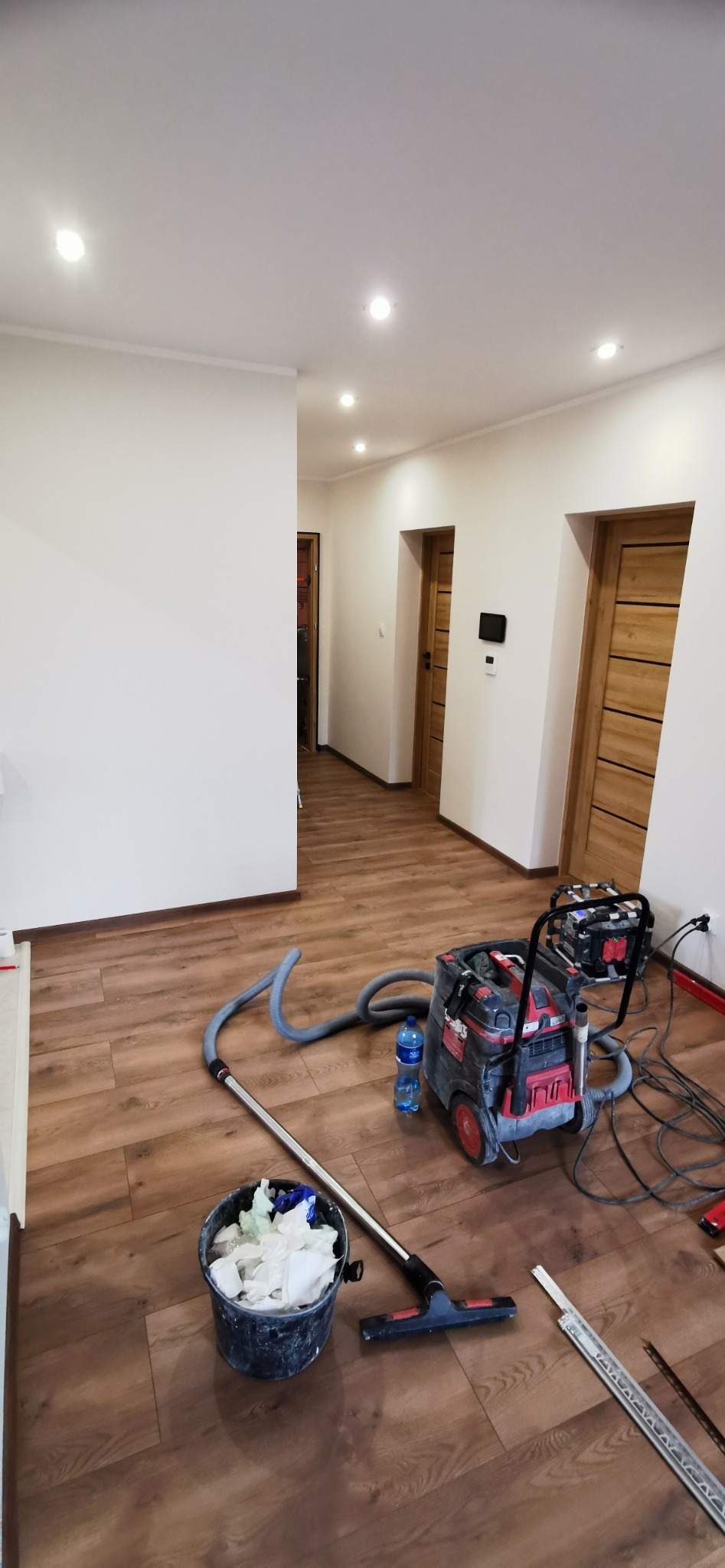 Korytarz po zakończonym układaniu paneli podłogowych, widoczny odkurzacz przemysłowy, wiadro z odpadami i listwy przypodłogowe do montażu.