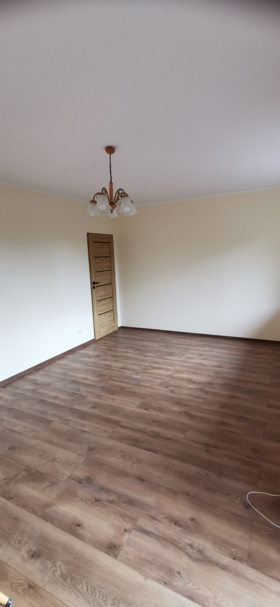 Puste pomieszczenie po remoncie z brązowymi panelami podłogowymi, beżowymi ścianami, drewnianymi drzwiami i wiszącym żyrandolem