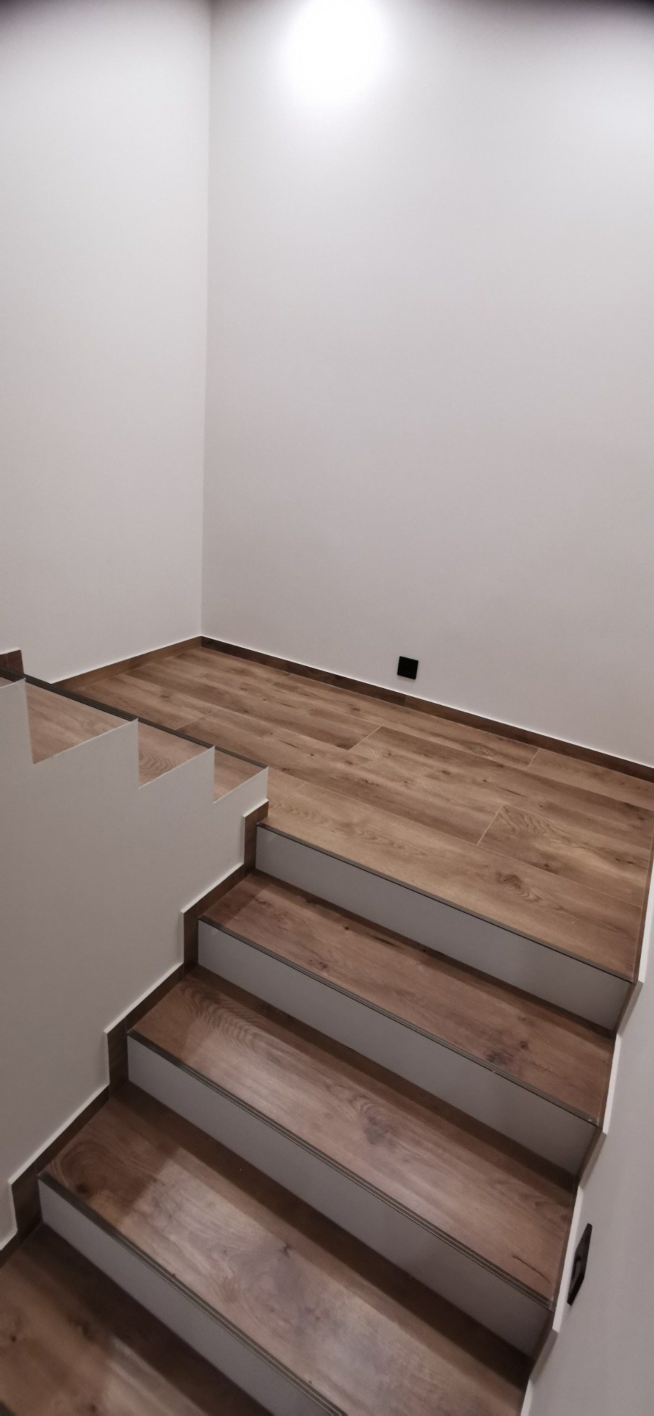 Schody wykończone panelami imitującymi drewno, z białymi podstopnicami i szarą listwą wykończeniową, widok z góry na dół, ściany białe z czarnym gniazdkiem elektrycznym.