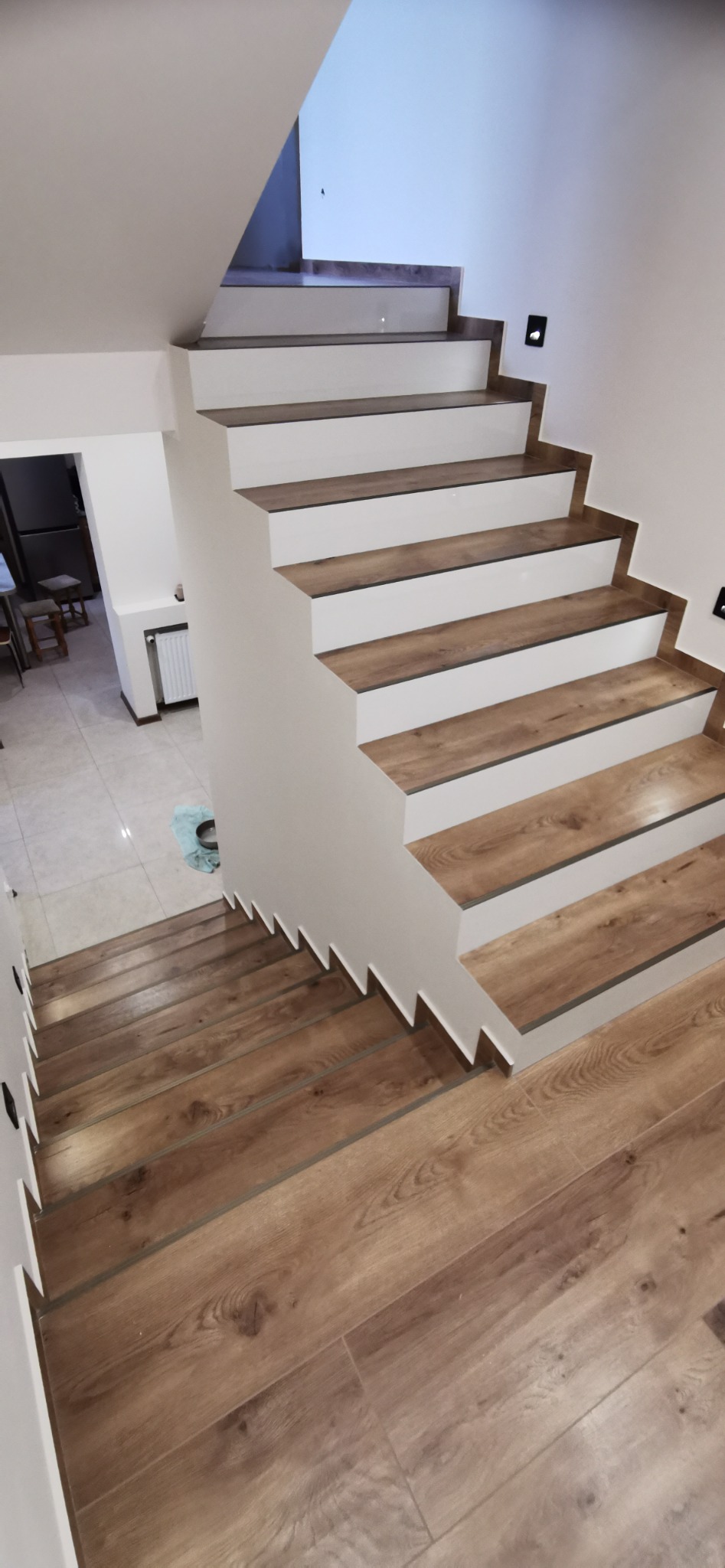 Nowoczesne schody z drewnopodobnymi panelami na stopniach i białymi podstopnicami, widok z góry, w tle fragment kuchni