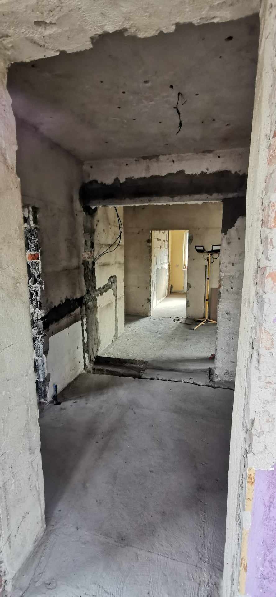 Wnętrze mieszkania w trakcie remontu: surowe ściany z widocznymi instalacjami elektrycznymi, betonowa podłoga i sufit, oświetlenie budowlane w tle.