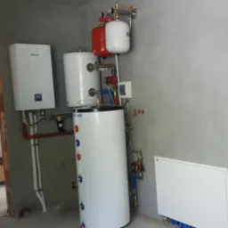 Biały zasobnik ciepłej wody użytkowej z czerwonymi i białymi zbiornikami wyrównawczymi, zamontowany w surowym pomieszczeniu z szarą ścianą i widocznymi elementami instalacji.