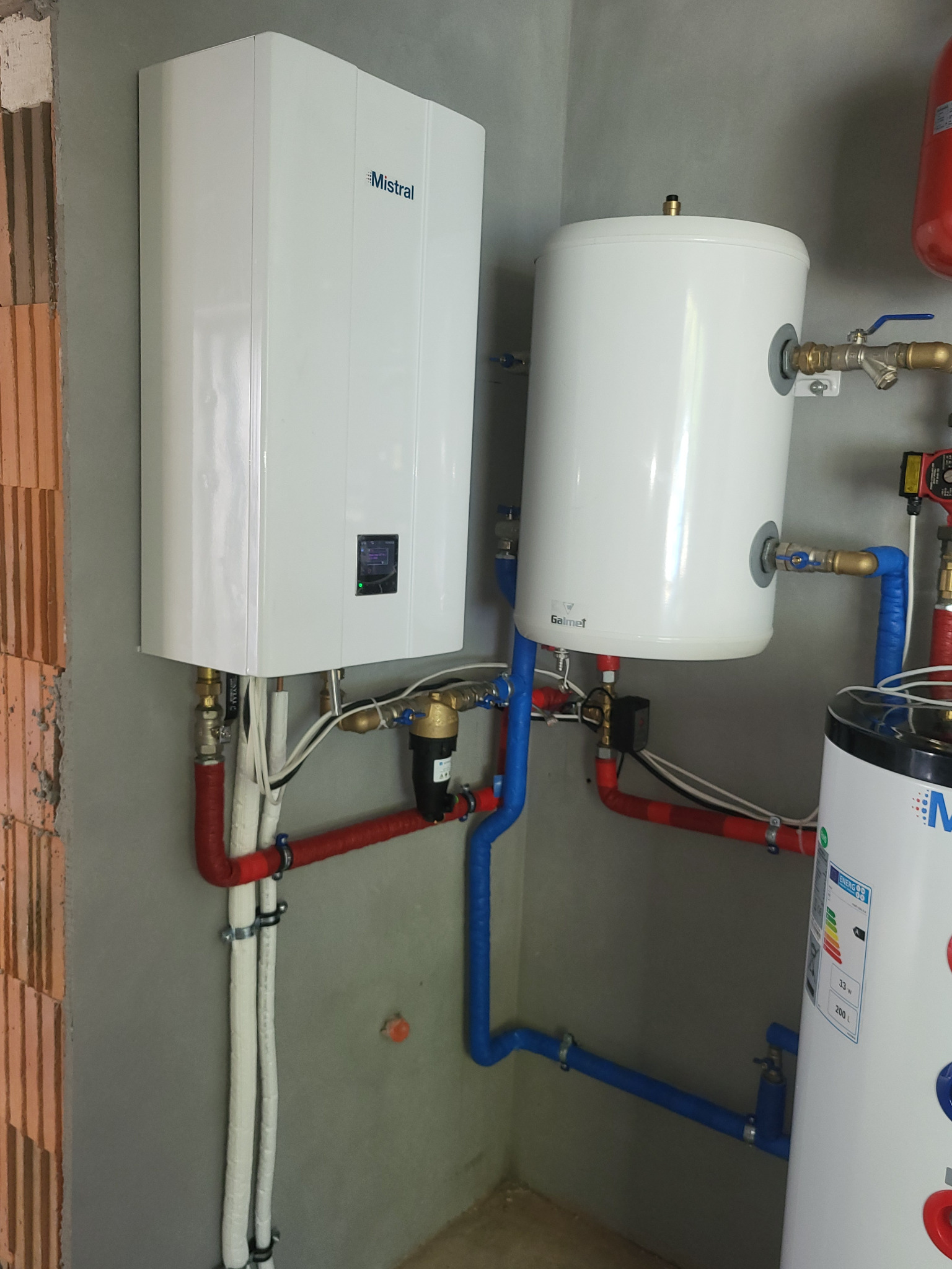 Biały, wiszący piec grzewczy Mistral z podłączonymi rurami miedzianymi i plastikowymi, obok zbiornik Galmet i fragment kolejnego zbiornika, montaż instalacji w surowym pomieszczeniu.