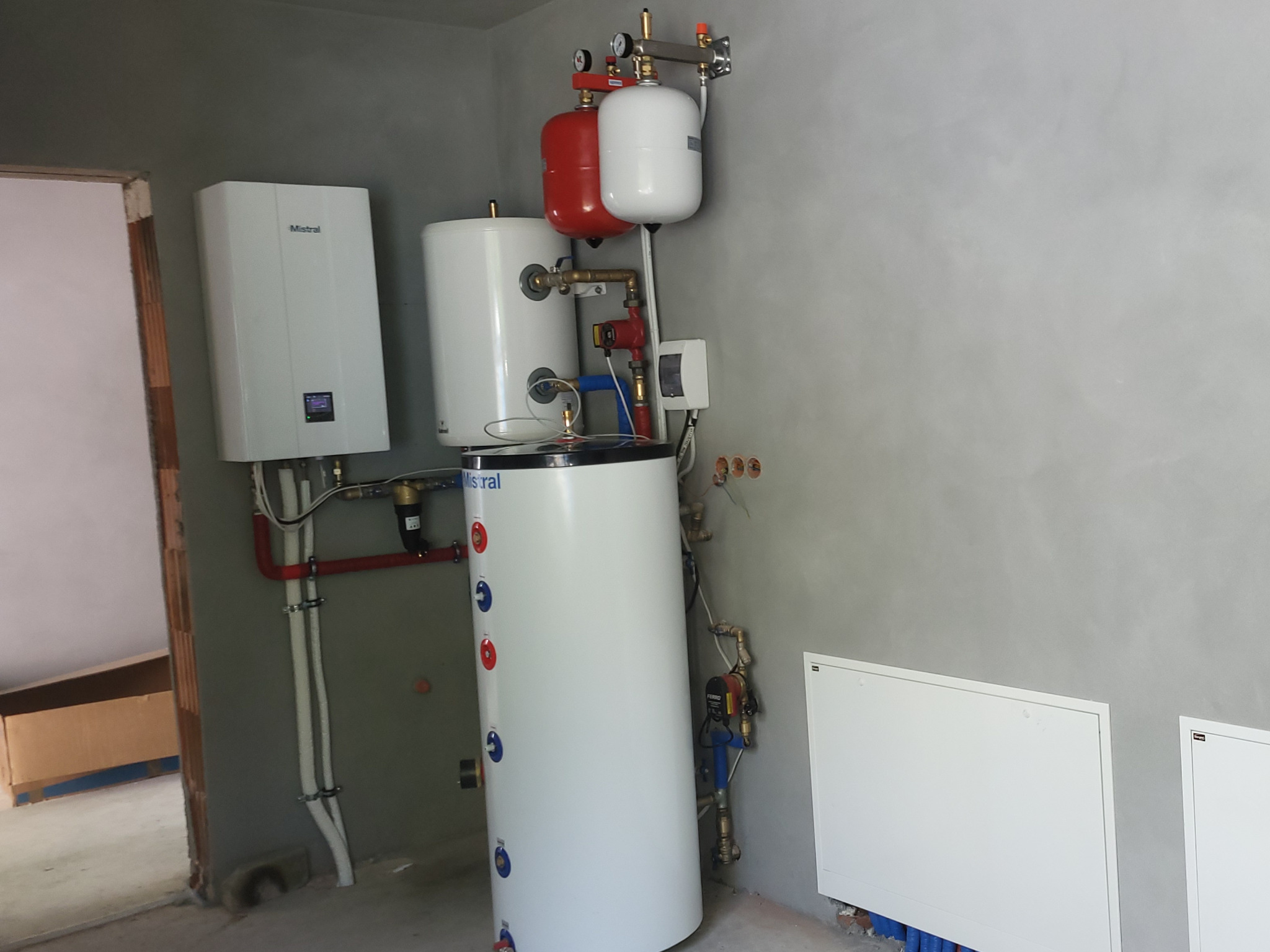 Biały zasobnik ciepłej wody użytkowej z czerwonymi i białymi zbiornikami wyrównawczymi, zamontowany w surowym pomieszczeniu z szarą ścianą i widocznymi elementami instalacji.