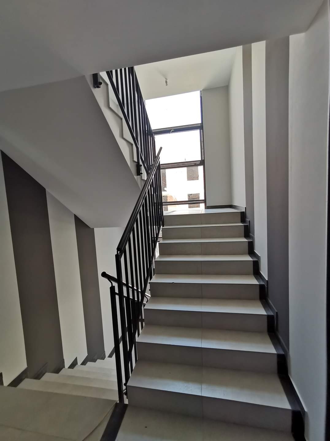 Klatka schodowa w budynku mieszkalnym, widok na schody z szarymi akcentami na ścianach i czarną balustradą, okno na końcu biegu schodów.