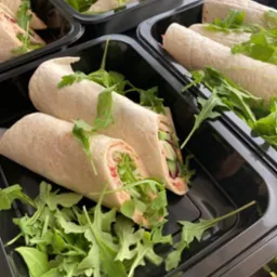 Wrapy i trójkątne kanapki z rukolą w czarnych pojemnikach na wynos, widoczne warstwy nadzienia, świeże i gotowe do spożycia.