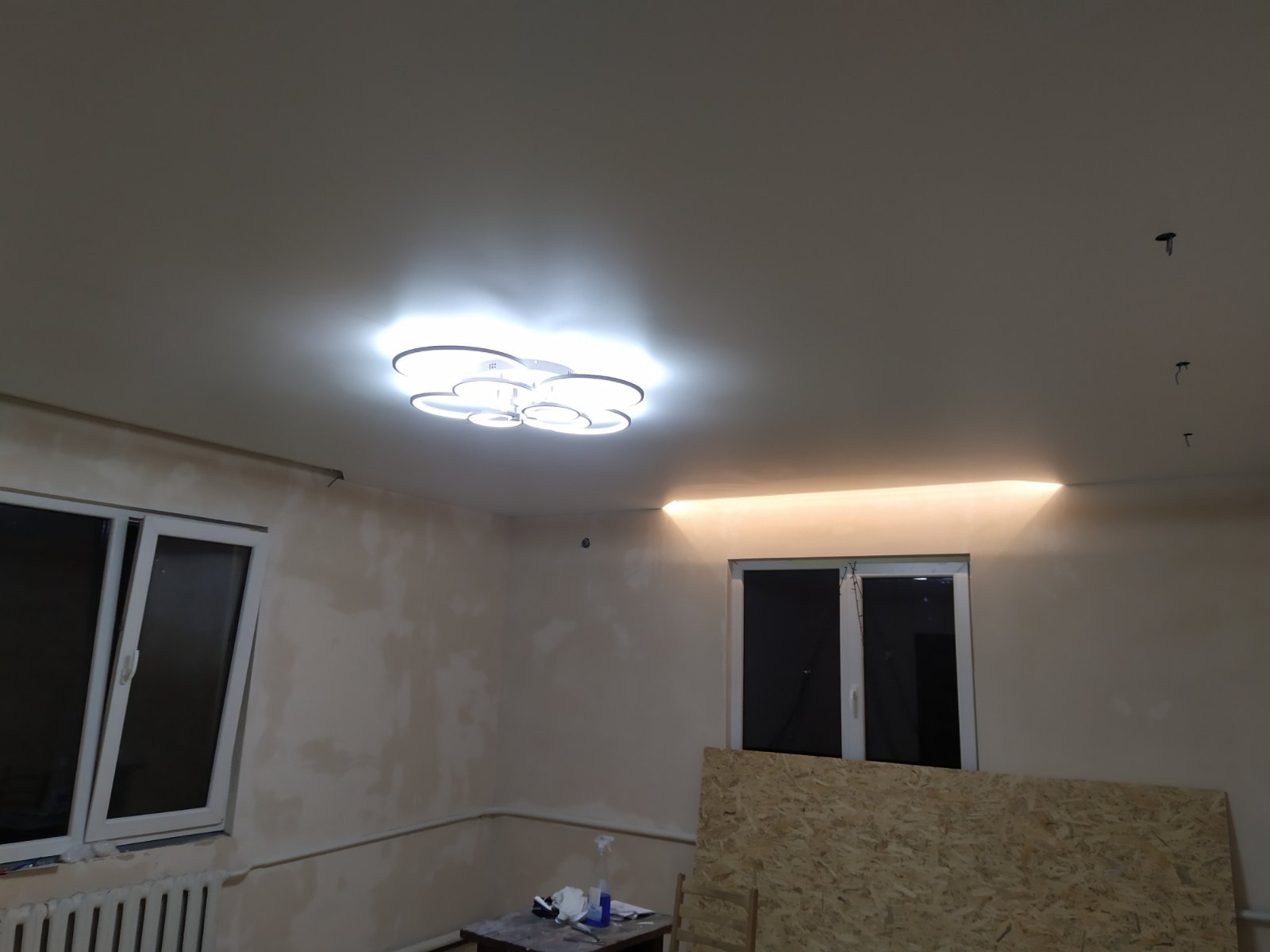 Pokój w trakcie prac wykończeniowych: szpachlowane ściany, sufit z oświetleniem LED i lampą w kształcie kwiatu, zabezpieczona płyta OSB przy oknie.