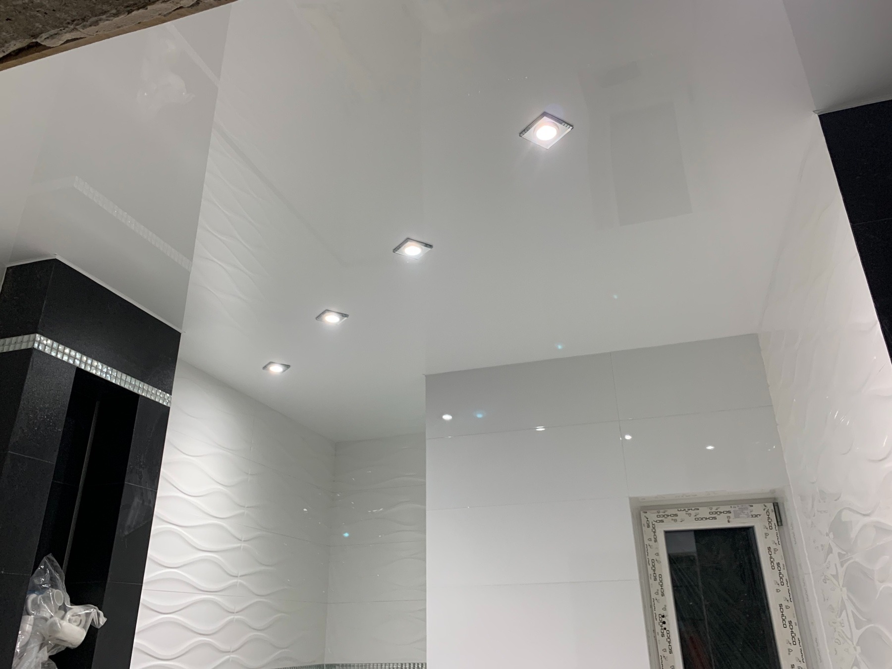 Biała, błyszcząca łazienka z podwieszanym sufitem i wbudowanymi, kwadratowymi lampami LED, oraz dekoracyjnymi płytkami ściennymi z motywem fal.