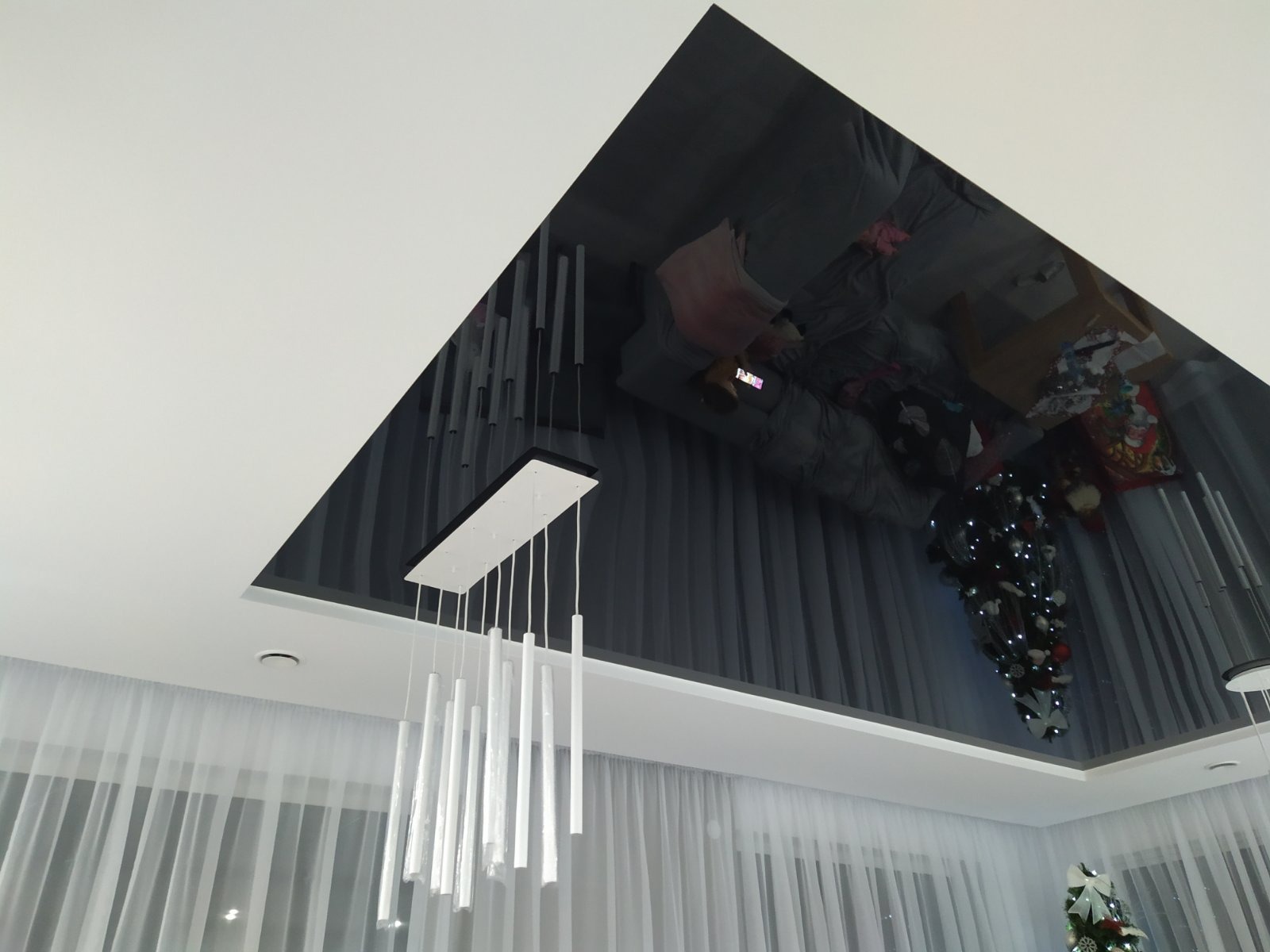 Czarny, lustrzany sufit napinany z odbiciem pokoju, oświetlenie LED w kształcie rurek, biały sufit i firanki.