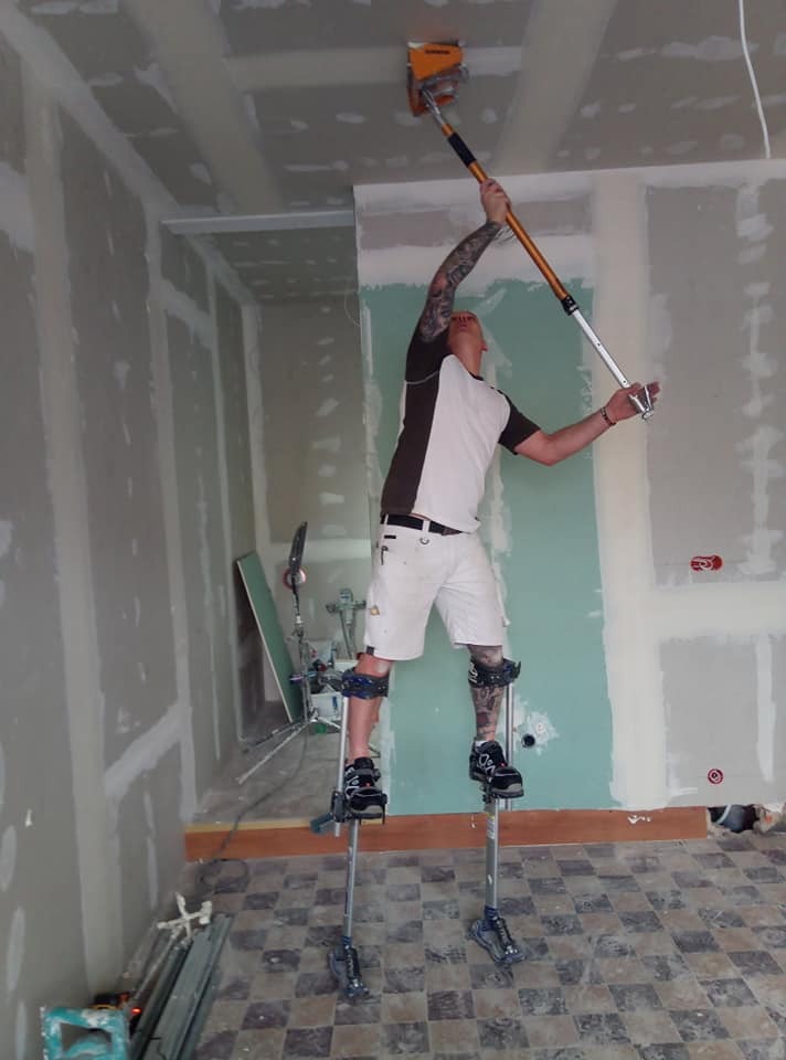Mężczyzna z tatuażami na szczudłach tynkuje sufit za pomocą narzędzia na długim wysięgniku w pomieszczeniu z płytami gipsowo-kartonowymi.