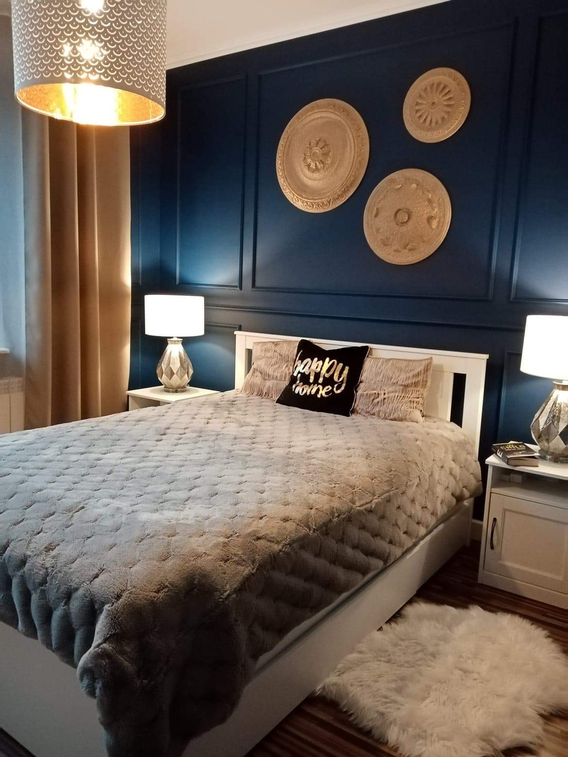 Elegancka sypialnia z granatowymi ścianami ozdobionymi dekoracyjnymi rozetami, białe meble, puszysty koc na łóżku i lampy nocne z dekoracyjnymi podstawami.