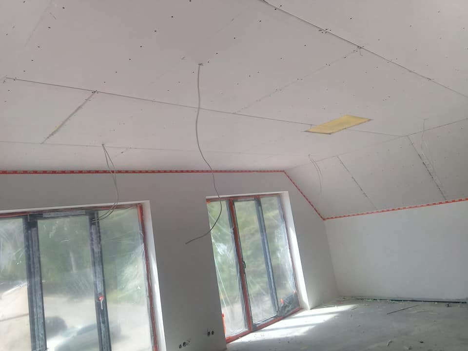Poddasze w trakcie prac wykończeniowych: ściany pomalowane na biało, okna zabezpieczone folią, sufit z płyt gipsowo-kartonowych ze śladami szpachlowania i wystającymi kablami.