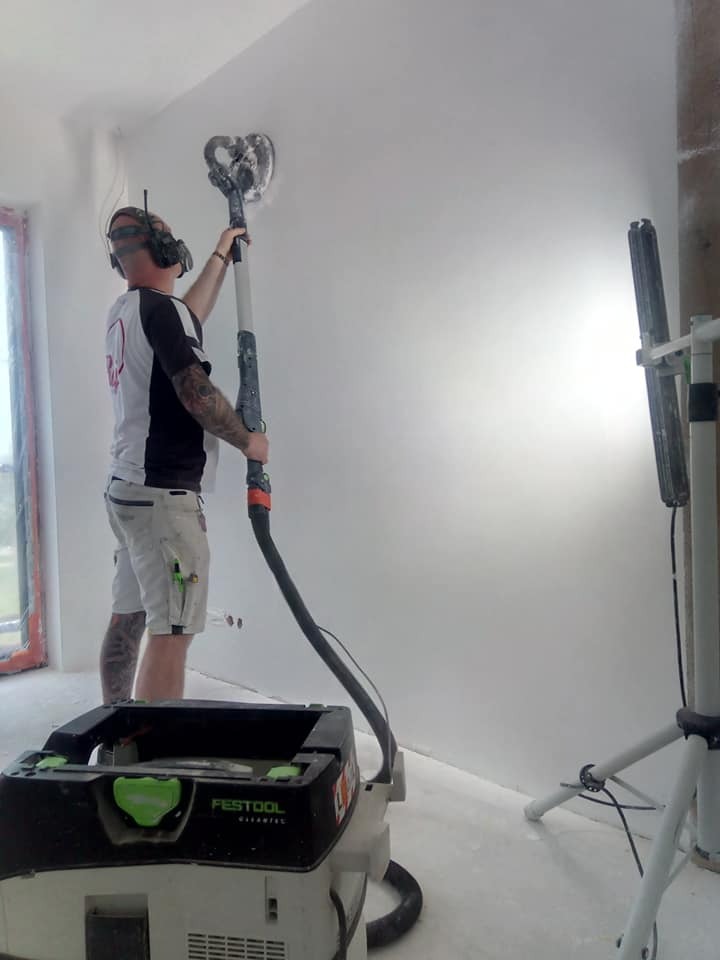 Pracownik w okularach ochronnych szlifuje ścianę za pomocą szlifierki z odkurzaczem Festool, widoczny kabel i oświetlenie halogenowe.