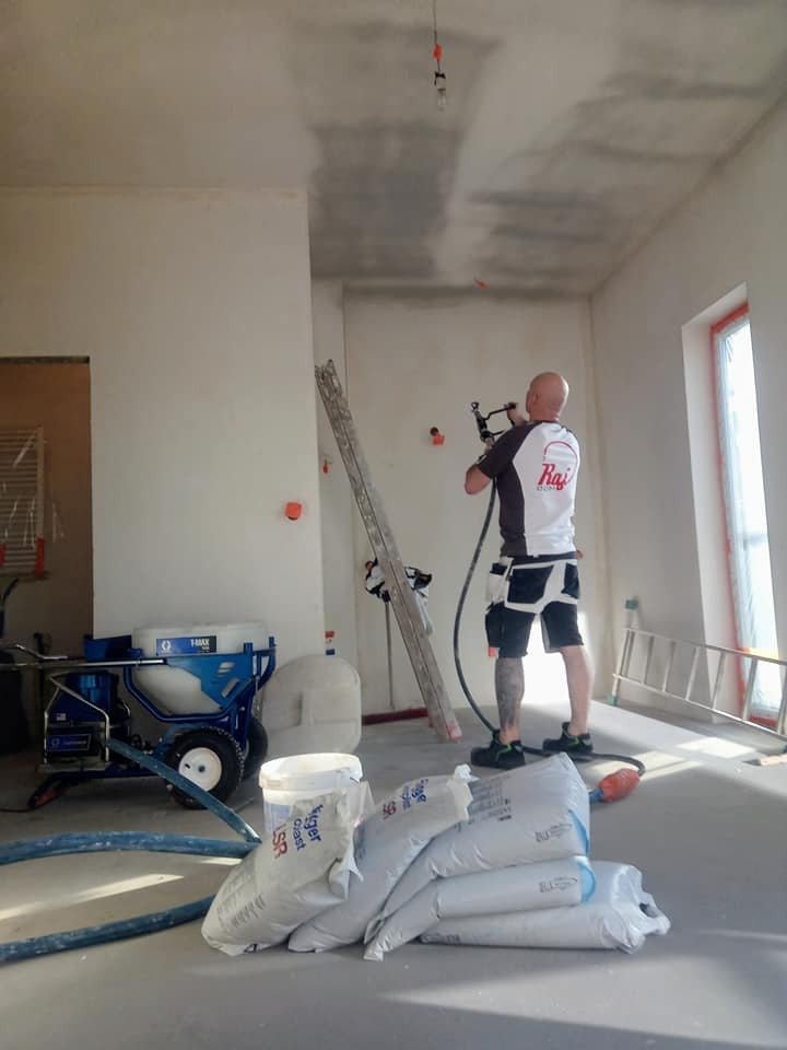 Pracownik w trakcie nakładania gładzi natryskowej na sufit w nowym budynku, widoczny agregat i worki z gładzią, surowe ściany i instalacje.