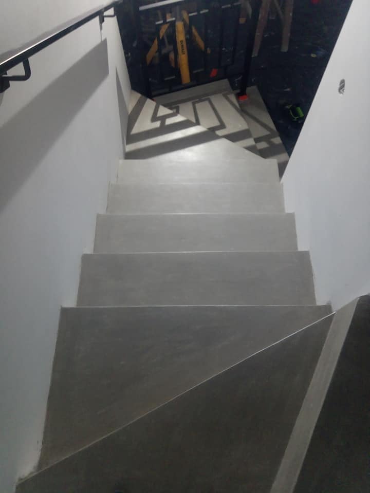 Widok z góry na pomalowane na szaro schody z czarną poręczą, na górze widoczny fragment podłogi z geometrycznym wzorem i narzędzia budowlane.