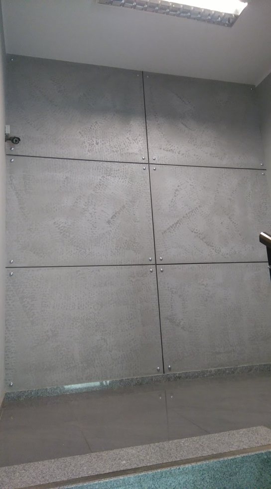 Ściana wykończona szarymi panelami z dekoracyjną fakturą, połączonymi czarnymi listwami i srebrnymi śrubami, oświetlona sztucznym światłem